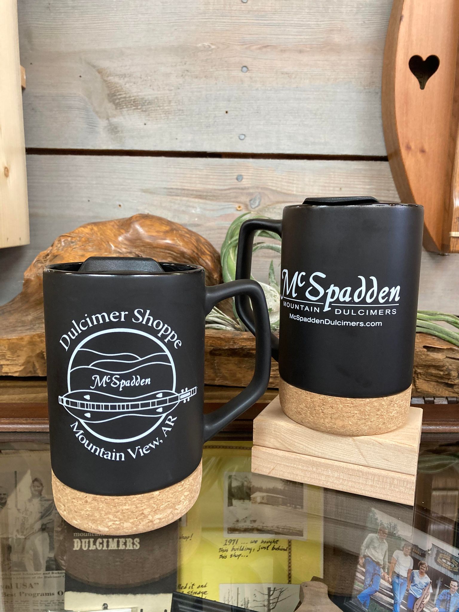 Two McSpadden Dulcimer Shoppe black coffee mugs with splash-proof lids on a shelf.