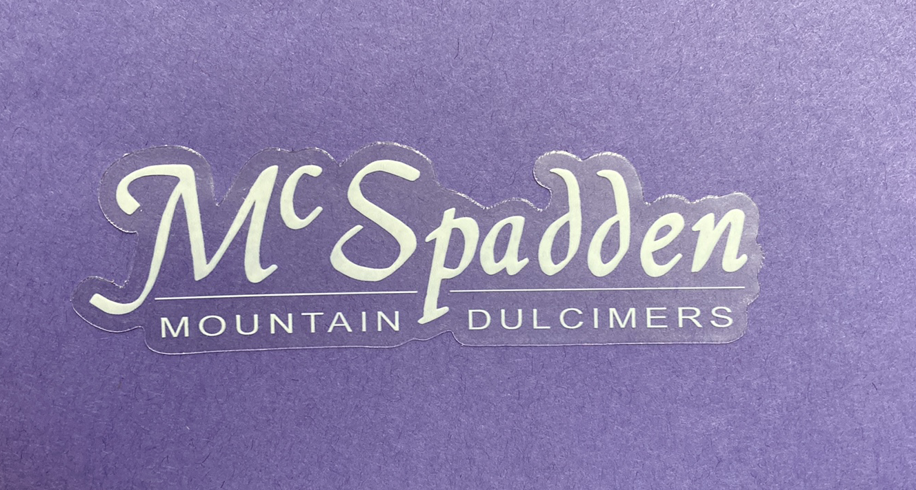 Sentence with product name: Silver logo of McSpadden mountain dulcimers decorating a purple background with McSpadden stickers.