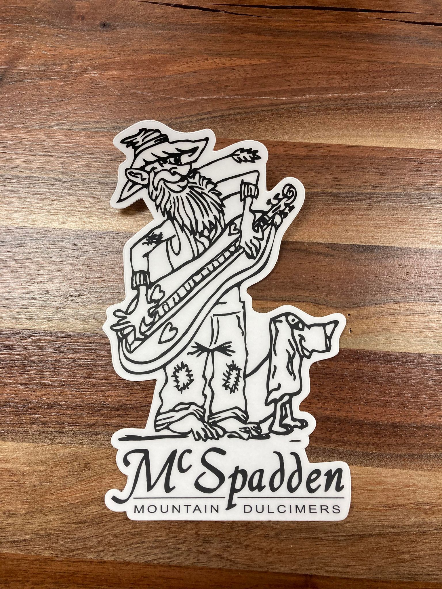 A McSpadden Sticker featuring an illustrated character playing a stringed instrument with a dog beside it, labeled "mcspadden mountain dulcimers," perfect for decorating laptops, on a wooden surface.