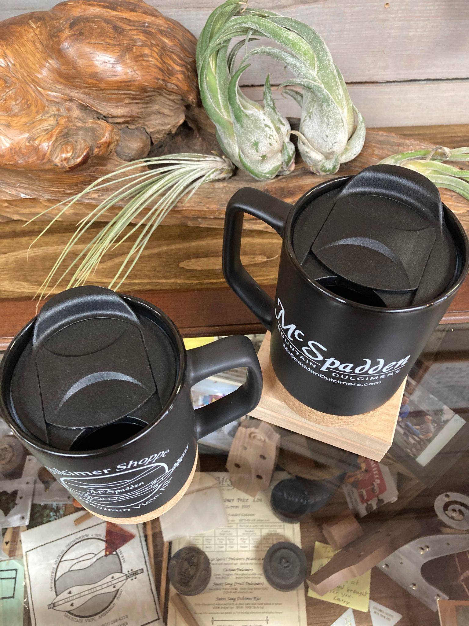 Two black McSpadden Dulcimer Shoppe travel mugs on a wooden stand.