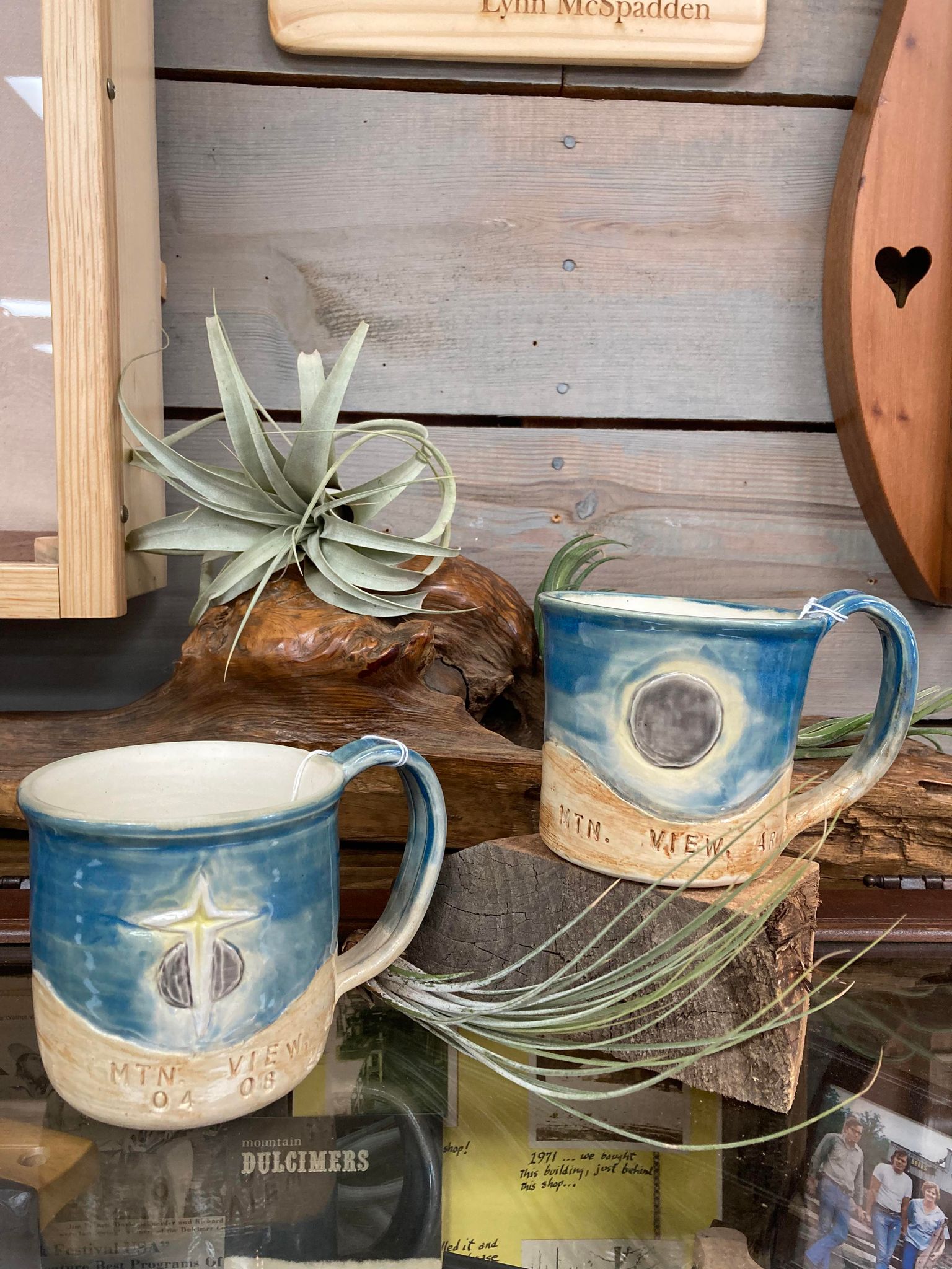 Handcrafted Total Solar Eclipse Pottery Mugs by Marion Ivey on a wooden shelf with artisanal design elements and a decorative air plant.