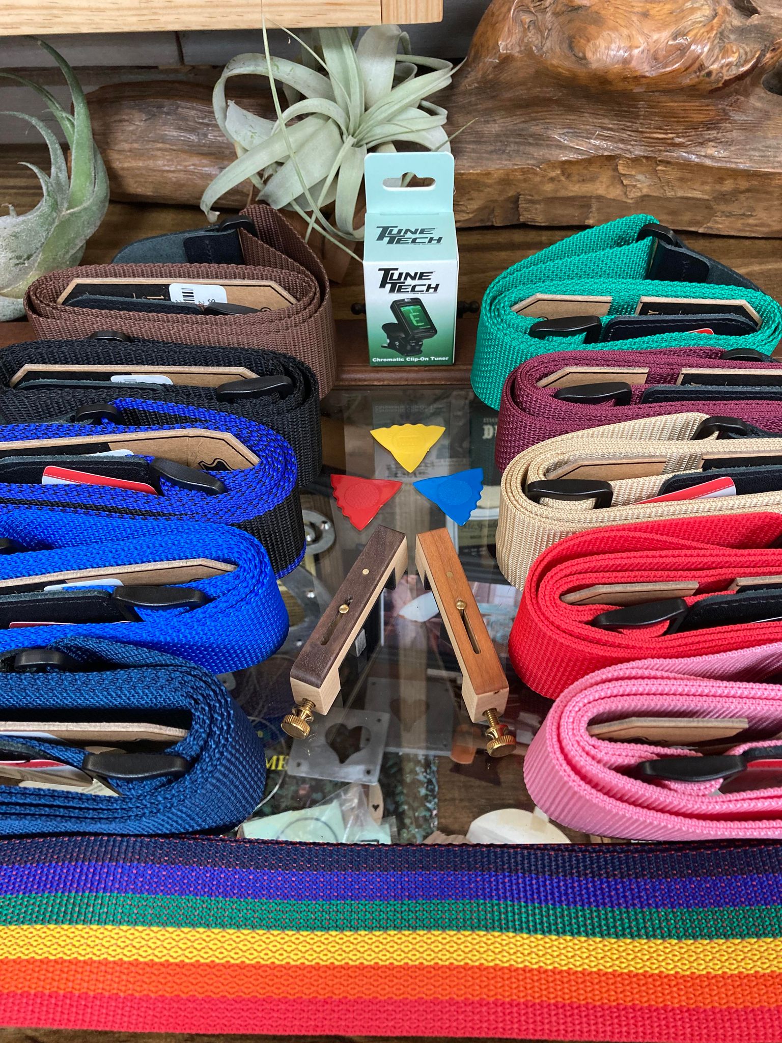 Colorful woven belts displayed for sale on a wooden shelf with a potted plant and the Dulcimer Essentials Package with Tuner in the background.