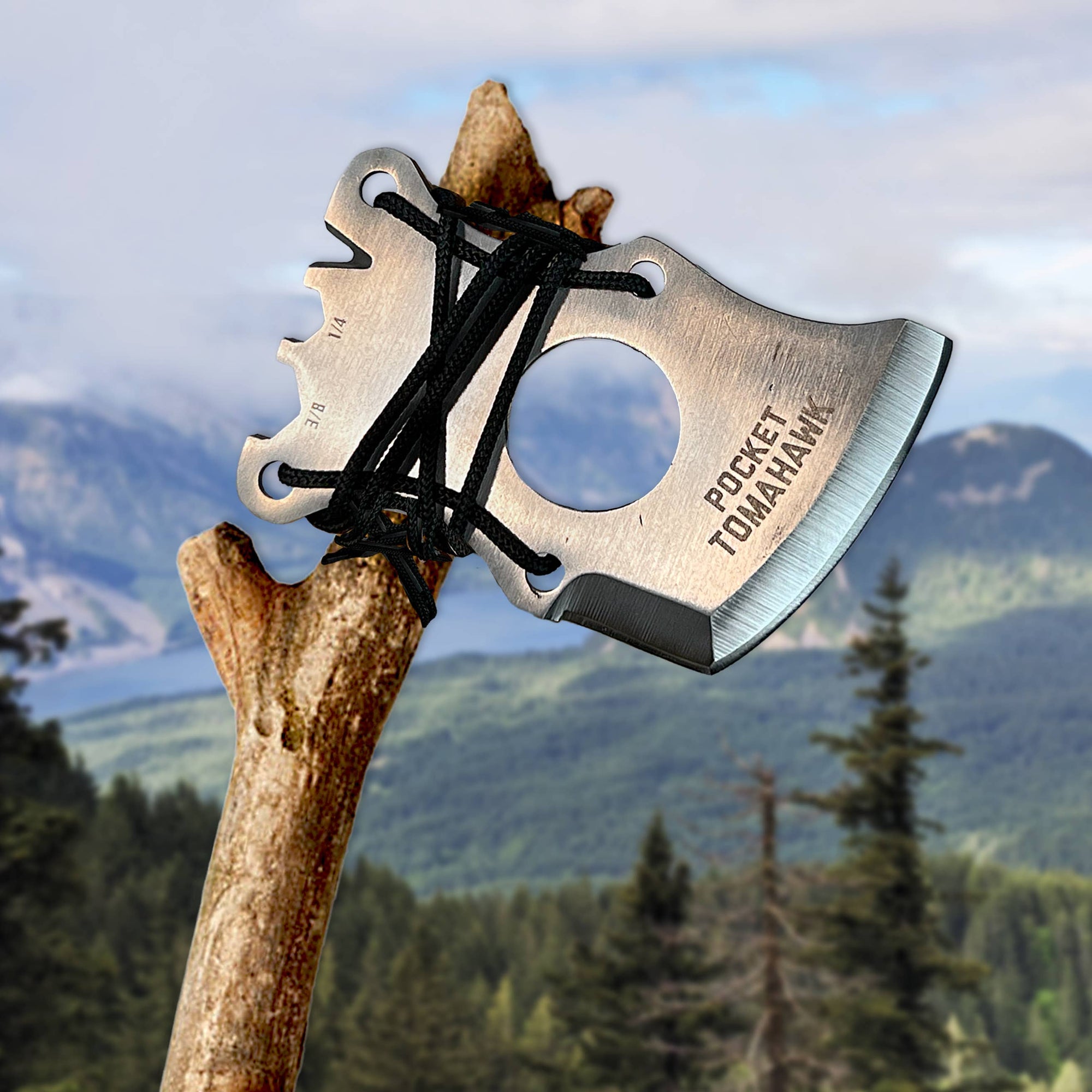 A close-up of a Pocket Tomahawk Axe Multi-Tool attached to a wooden handle with black 550 paracord rope, set against a blurred mountain landscape.