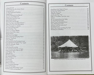 Black and white photo of an outdoor gospel music event with people seated under a large tent in a field, the event setup visible in a countryside setting. Adjacent page shows a table of contents of "Hymns of the Old Camp Ground" by Wayne Erbsen on old time gospel songs history.