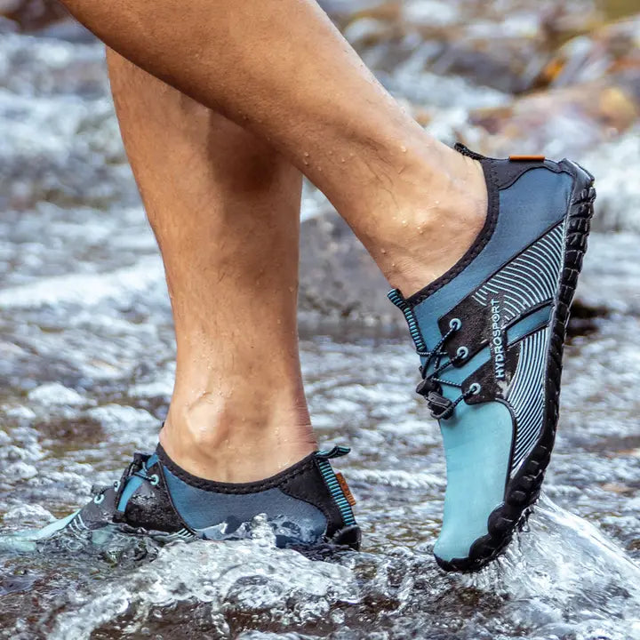 A pair of feet adorned in blue and black HydroSport FitKicks, stepping through a shallow, rocky stream.