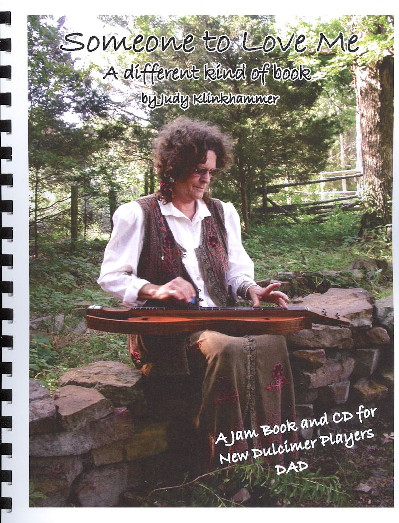 Person playing a dulcimer outdoors on a stone bench in a forested setting, with an instruction book titled "Someone To Love Me (D-A-D) - by Judy Klinkhammer displayed