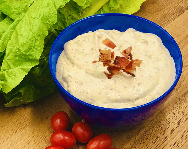 A bowl of BLT Dip Mix topped with crispy bacon pieces, accompanied by lettuce leaves and cherry tomatoes on a wooden surface.