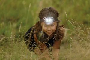 A young girl wearing an Outdoor Discovery Critter Head Lamp crawls through tall grass, focusing intently ahead.