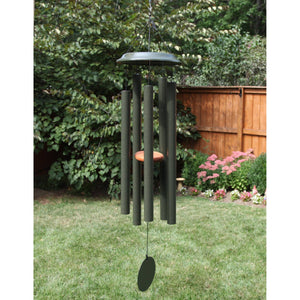 The 47-inch Shenandoah Melodies® green wind chime, hand-tuned with multiple tubes, becomes a striking attraction, hanging gracefully in a backyard with grass, a wooden fence, and bushes.