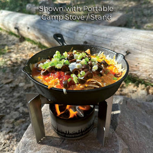 A skillet of chili topped with cheese and scallions, cooking over a City Bonfires® by Coleman®, set outdoors on a wooden surface.