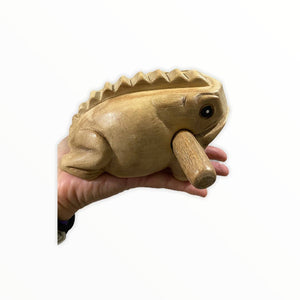 A hand holding a Natural wooden frog, handmade in Thailand, with a mallet in its mouth. It’s used for creating sound by scraping, showcasing quality craftsmanship throughout.