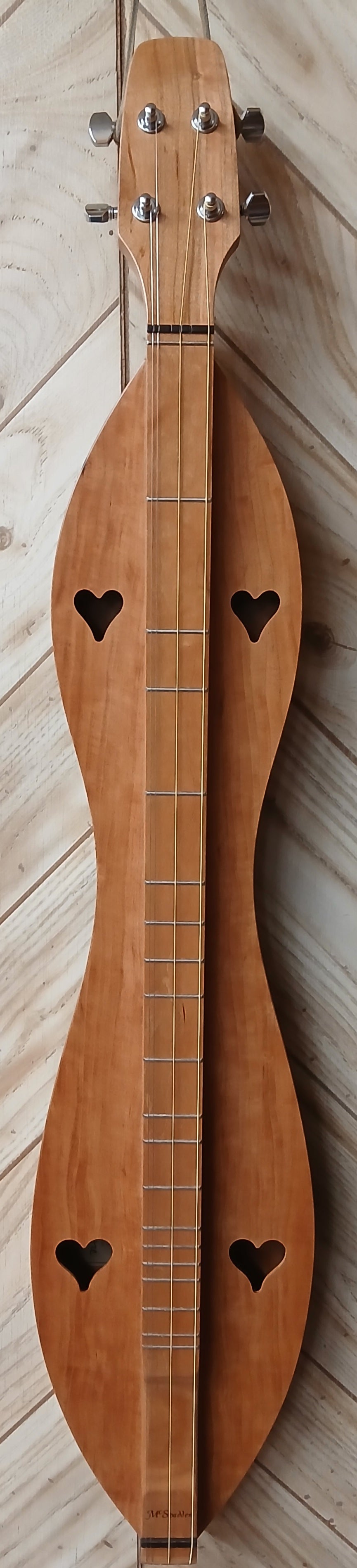 The handcrafted 4FHCCB, a teardrop-shaped wooden string instrument with four tuning pegs and two heart-shaped sound holes, rests on the floor. It's a customizable dulcimer perfect for any musician looking to add an artistic touch to their collection.