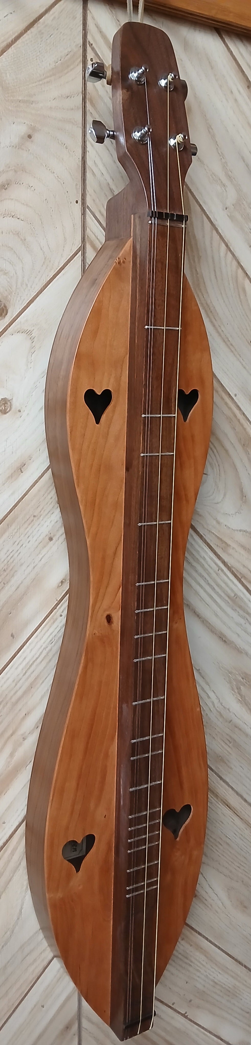 A 4 String Baritone dulcimer, with an hourglass design and walnut back and sides, featuring a cherry top and heart-shaped sound holes, leans against a light wood-paneled wall. (Model: 4FHWCB).