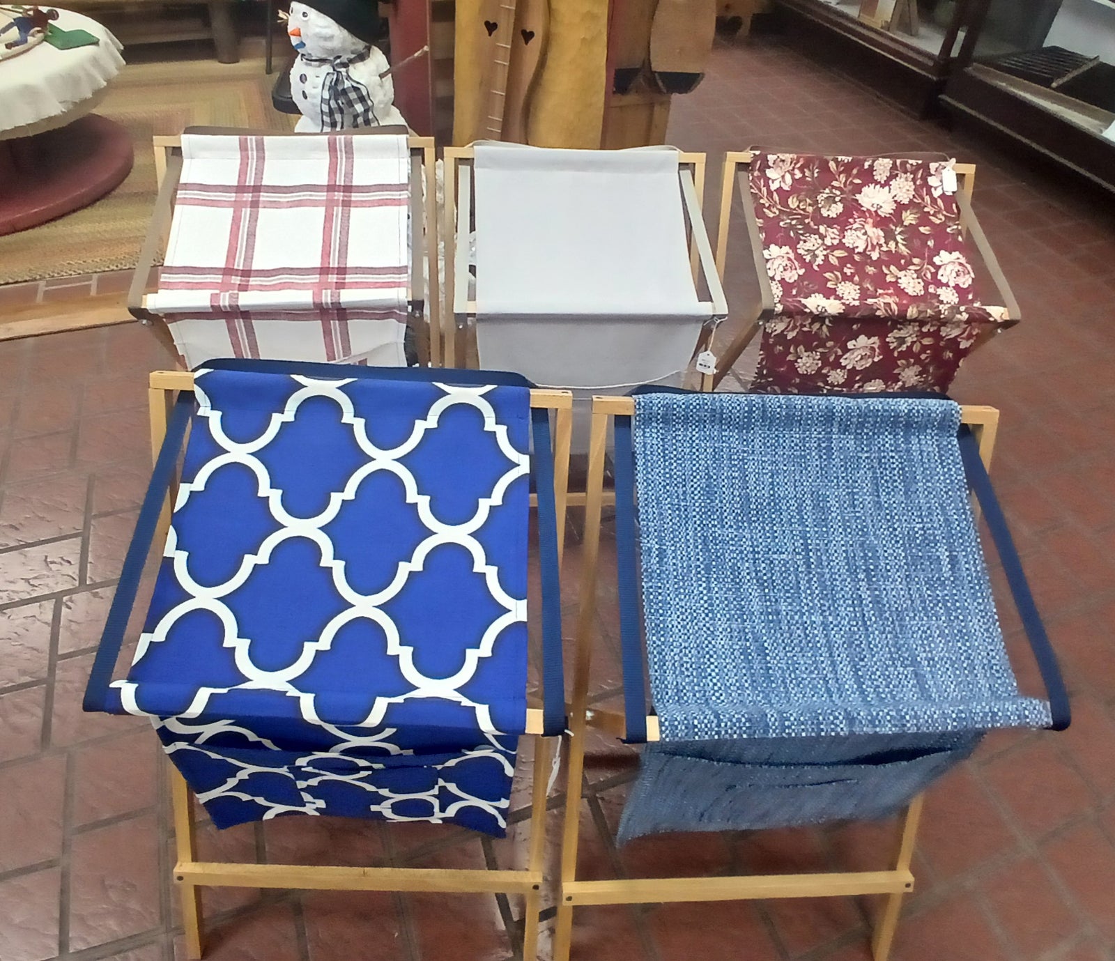 Five Sling Music Stand hampers with various patterned fabric liners are displayed in two rows on a brick floor inside a shop.