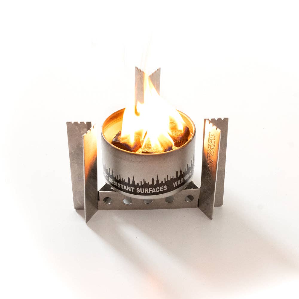 A Portable Camp Stove / Stand Combo with a visible flame, encased in a metal frame with safety warnings on the side, against a white background.