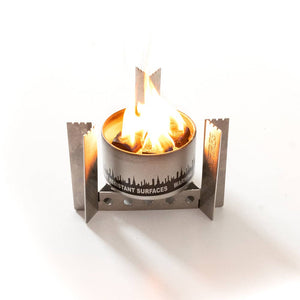 A Portable Camp Stove / Stand Combo with a visible flame, encased in a metal frame with safety warnings on the side, against a white background.