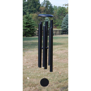 The Corinthian Bells® 65-inch Windchime - Wholesale, a large black metal chime with four tubes and deep resonance, hangs outdoors in a grassy yard with trees and a wooden fence in the background.