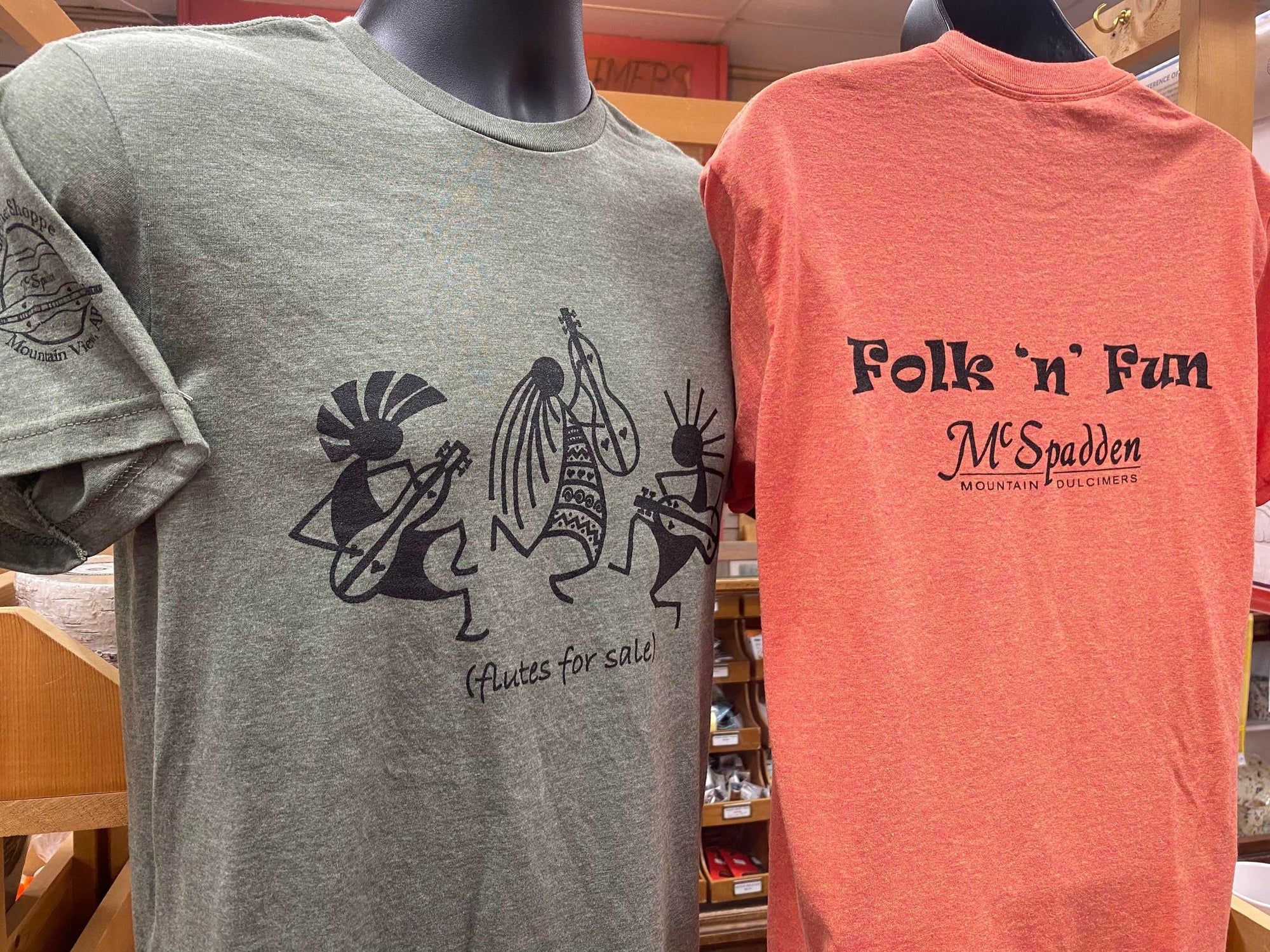 Two t-shirts on display: one gray with a design of people playing flutes and text "Flutes for Sale," and one orange featuring a design of Kokopelli playing dulcimers with text "Folk 'n' Fun McSpadden Mountain Dulcimers.