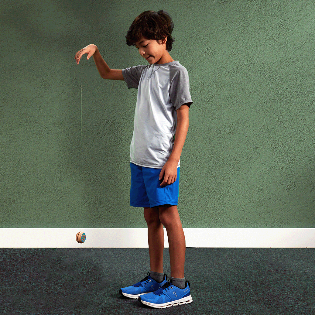 A young boy playing with a Neato! wood yo-yo against a green wall, wearing a gray t-shirt, blue shorts, and blue sneakers.