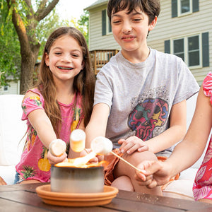 Two children smiling while roasting marshmallows over a Mini Portable Fire Pits - City Bonfires® by Coleman® outdoors.