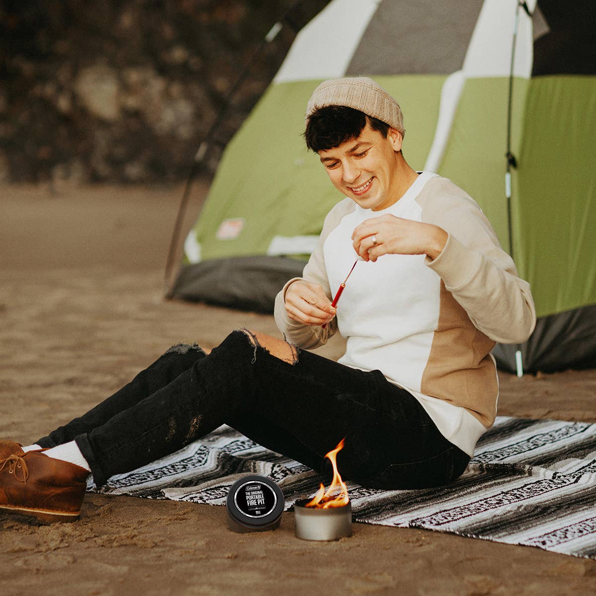 A person sitting on a rug near a tent, smiling while lighting a City Bonfires® by Coleman® with a match.