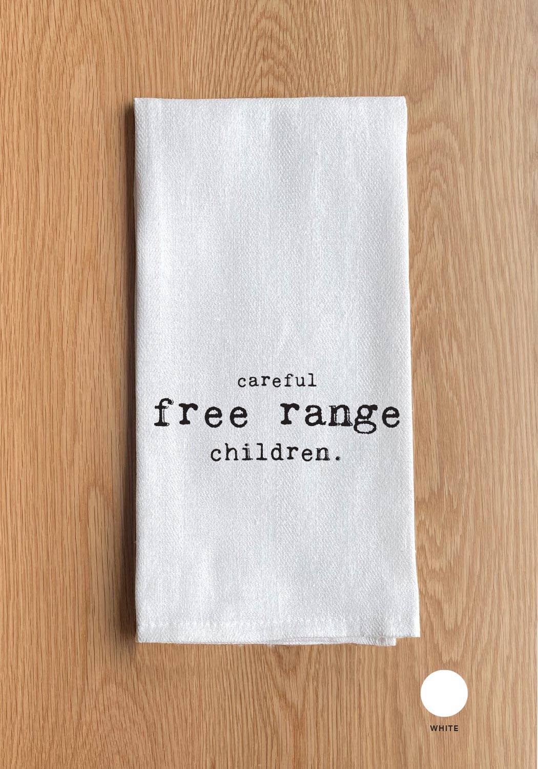 A 100% cotton towel featuring the printed words "Careful, Free Range Children" lies folded on a wooden surface.