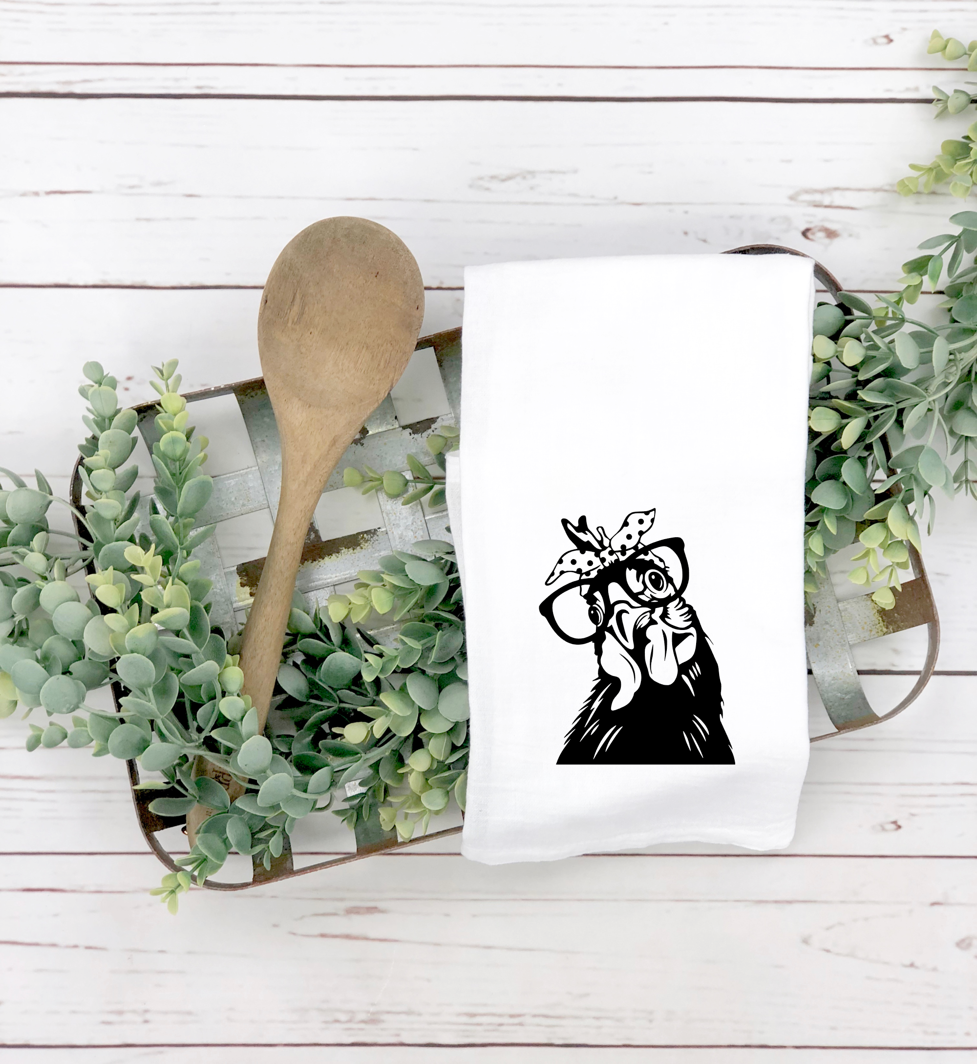 A Funny Chicken Tea Towel with a printed image of a chicken wearing glasses and a bow tie, accompanied by a wooden spoon, on a rustic white wooden background flanked by greenery.