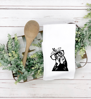 A Funny Chicken Tea Towel with a printed image of a chicken wearing glasses and a bow tie, accompanied by a wooden spoon, on a rustic white wooden background flanked by greenery.