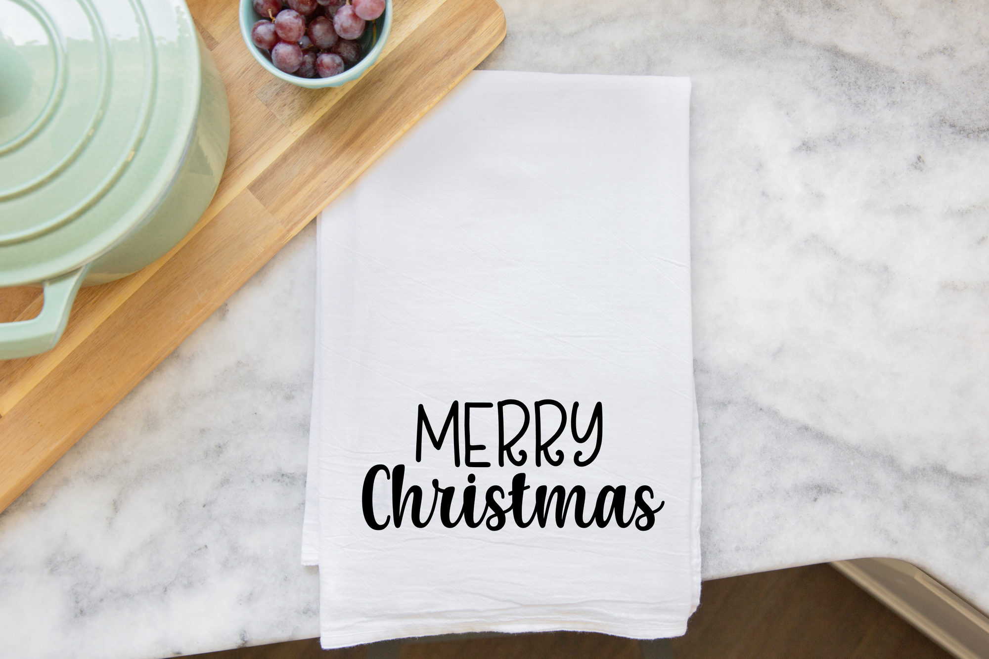 A Merry Christmas Tea Towel featuring a white background and black lettering is elegantly placed next to a wooden cutting board with grapes and a light green pot, creating a charming and affordable Christmas gift for any kitchen.