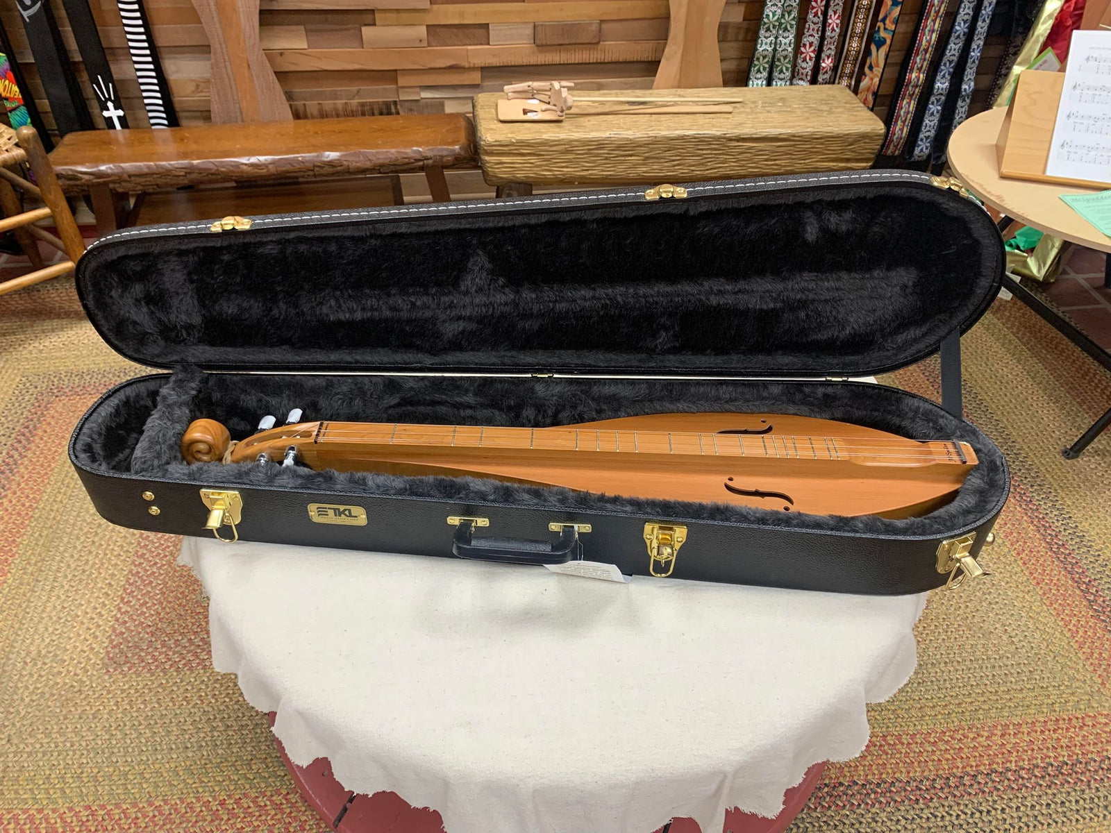 A mountain dulcimer sits in an open Premier Dulcimer Hard Case—black, velvet-lined hardwood with custom neck support—on a round, white-clothed table.