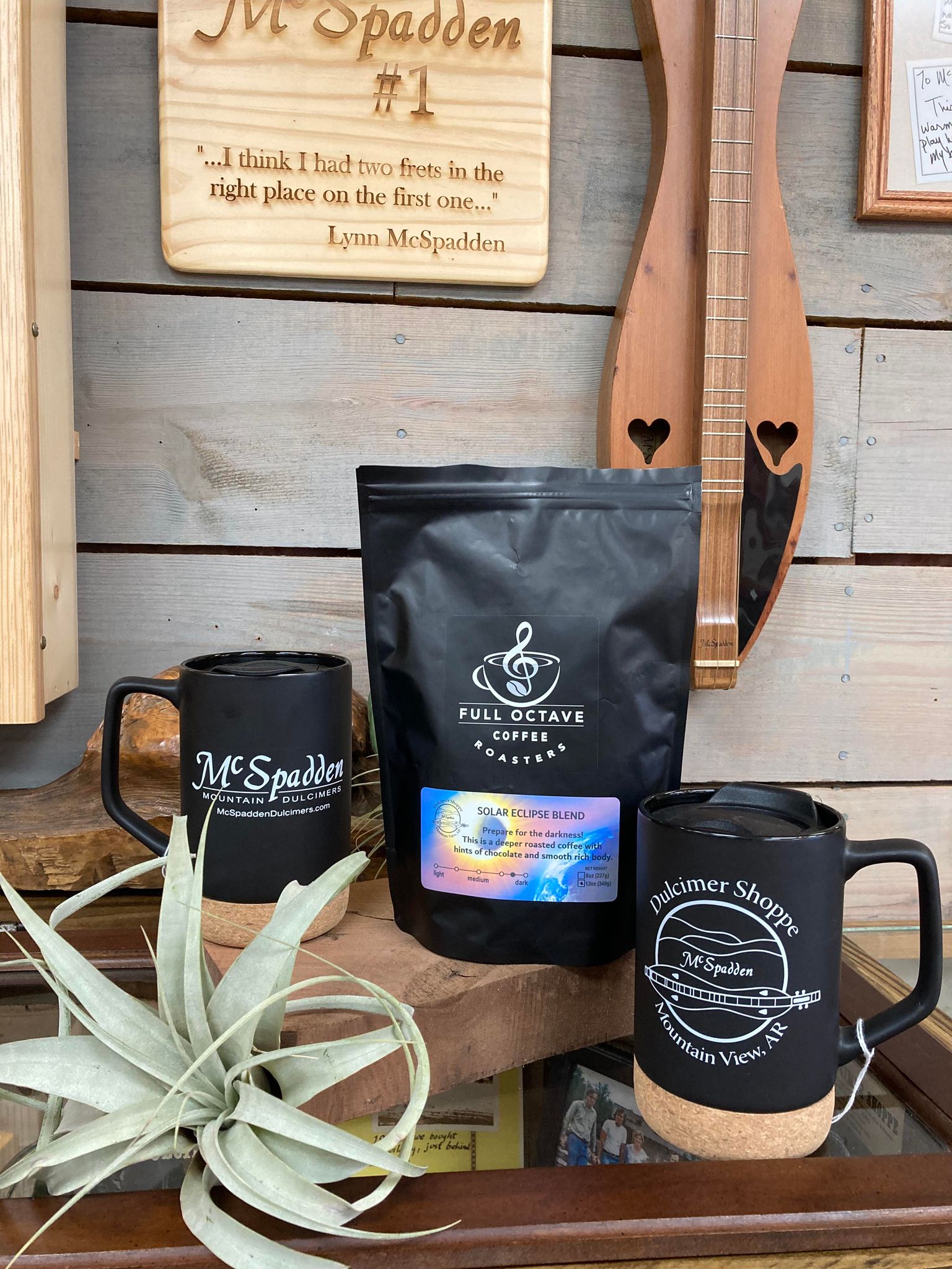 A display featuring a Full Octave Coffee bag, a wooden instrument, two McSpadden branded mugs, a potted plant, and a wall plaque with an engraved quote. Among the items is Full Octave Coffee medium roast coffee adding to the inviting ambiance.