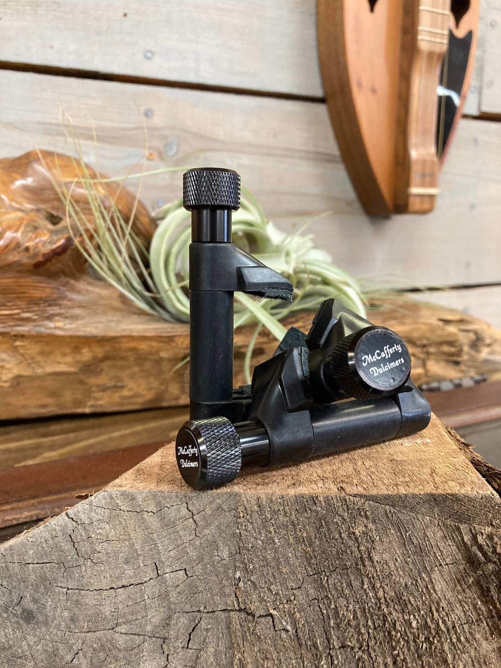 A black McCafferty Capo, crafted for six-string models, rests gracefully on a wooden surface against a rustic, wood-paneled backdrop. Its sleek design is enhanced by silicone padding, ensuring both functionality and style.