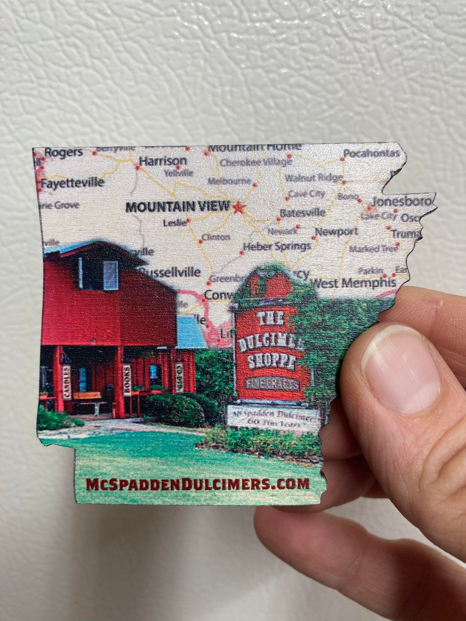 A hand holding an Arkansas Wooden Magnet shaped like the state, with a map backdrop and an image of The Dulcimer Shoppe in Mountain View. The text "MCSPADDENDULCIMERS.COM" is at the bottom, making it a perfect souvenir.