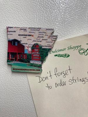 A charming Arkansas Wooden Magnet designed as a souvenir, featuring "The Dulcimer Shoppe" and a red building image, holding a note that reads, "Don't forget to order strings!".