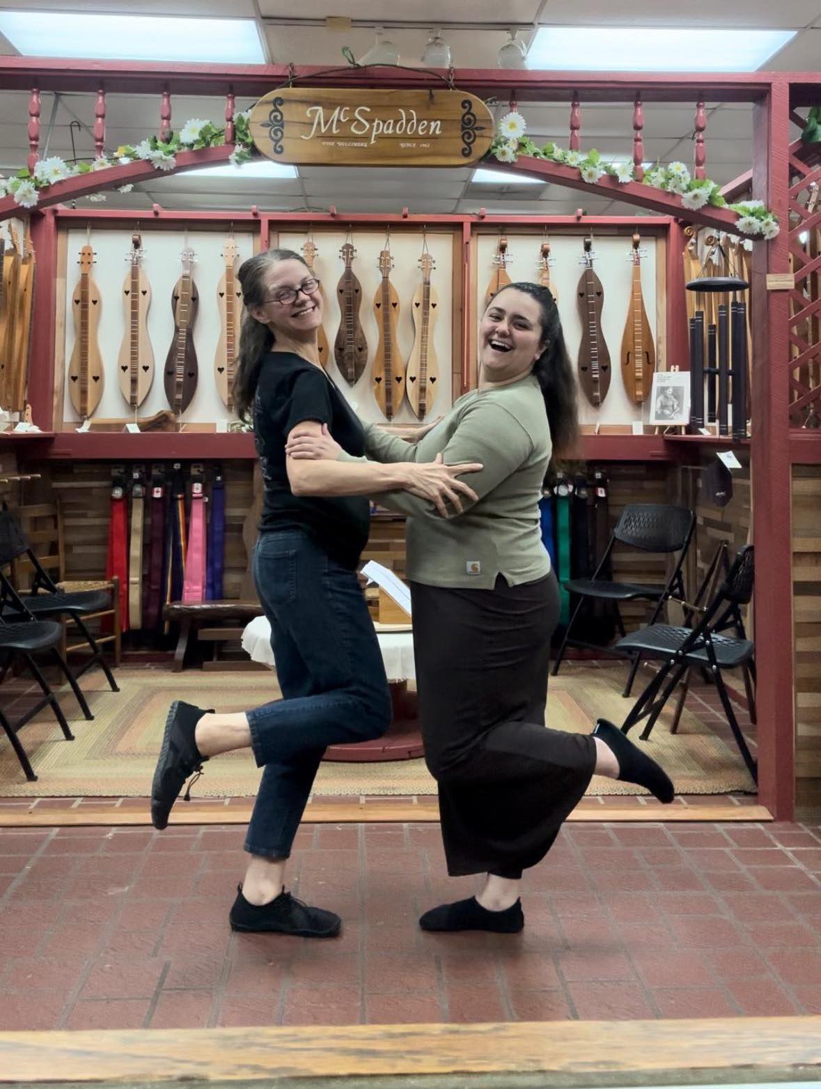 Two women smiling and dancing in front of a display of HydroSport FitKicks at McSpadden store, clasping hands and kicking up their heels in their amphibious footwear.