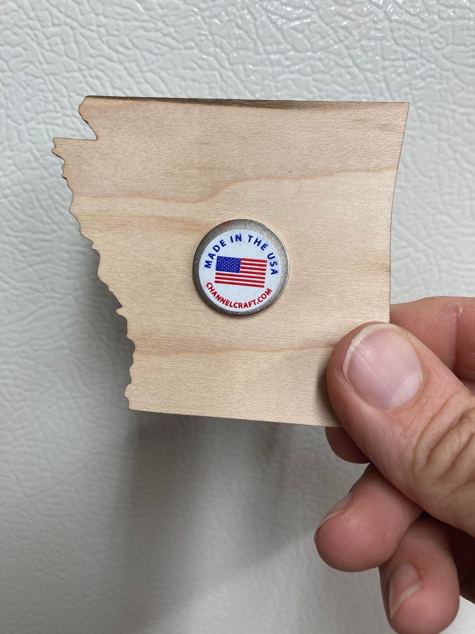 A hand holds a wooden cutout of a US state with a "Made in the USA" sticker featuring an American flag. This Arkansas Wooden Magnet from The Dulcimer Shoppe makes the perfect souvenir.