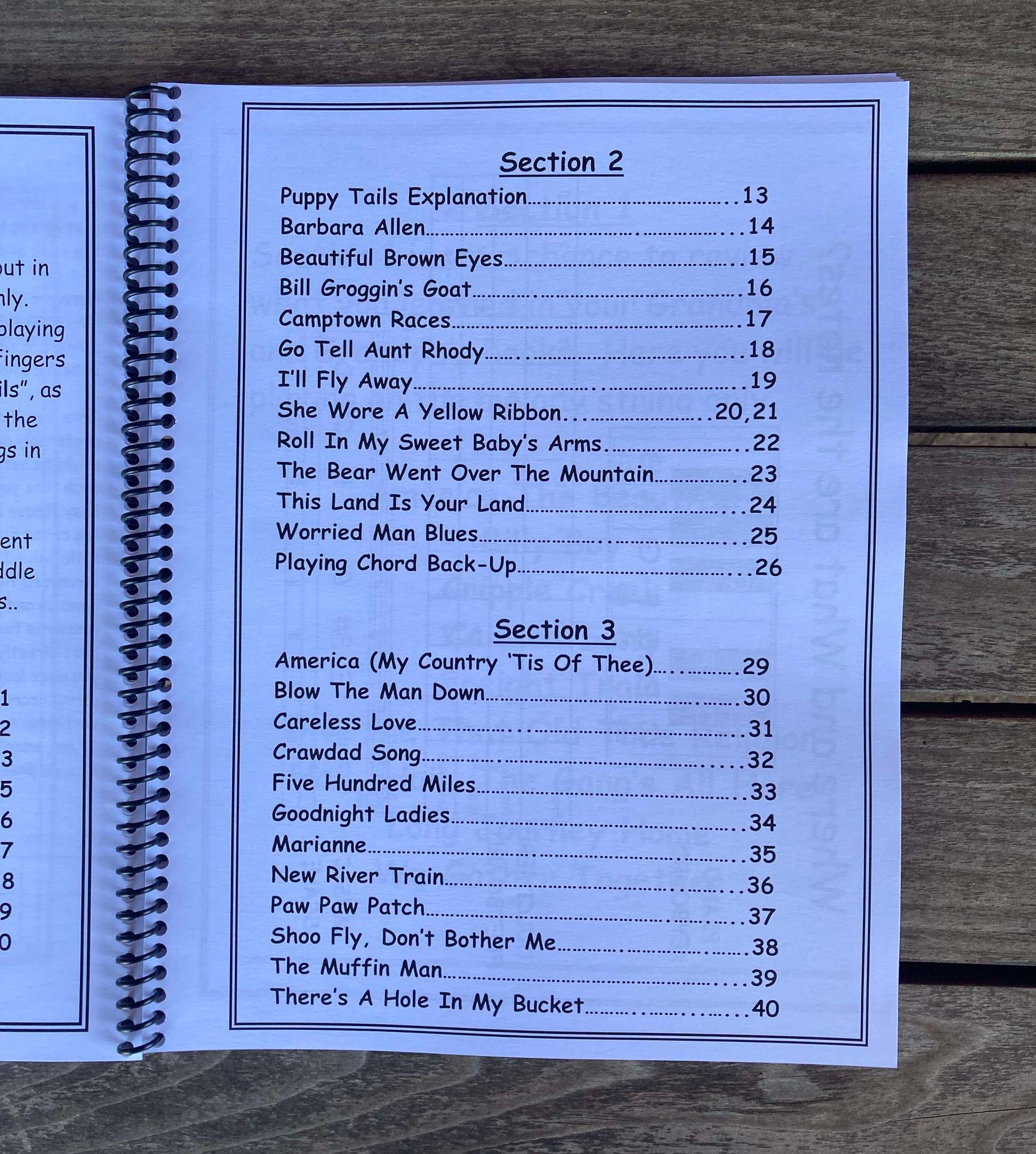 A spiral-bound intermediate book, Aunt Rhody's Step Up - by Red Dog Jam, lies open to a table of contents page listing happy music under Sections 2 and 3, including "She Wore A Yellow Ribbon" and "America (My Country 'Tis Of Thee)." The folk songs page is on a wooden surface.