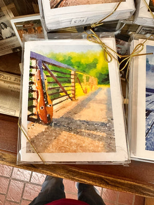 A watercolor painting by Ellen Pratt depicts a guitar leaning against a railing on a sunlit bridge, surrounded by greenery. The painting, stacked with other similar artworks tied with golden strings, exudes the charm often found in Dulcimer Greeting Card Sets by Ellen Pratt.