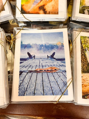 A printed image of a lakeside dock with two empty chairs, packaged in a clear plastic sleeve and tied with gold string for display, reminiscent of Dulcimer Greeting Card Sets by Ellen Pratt.