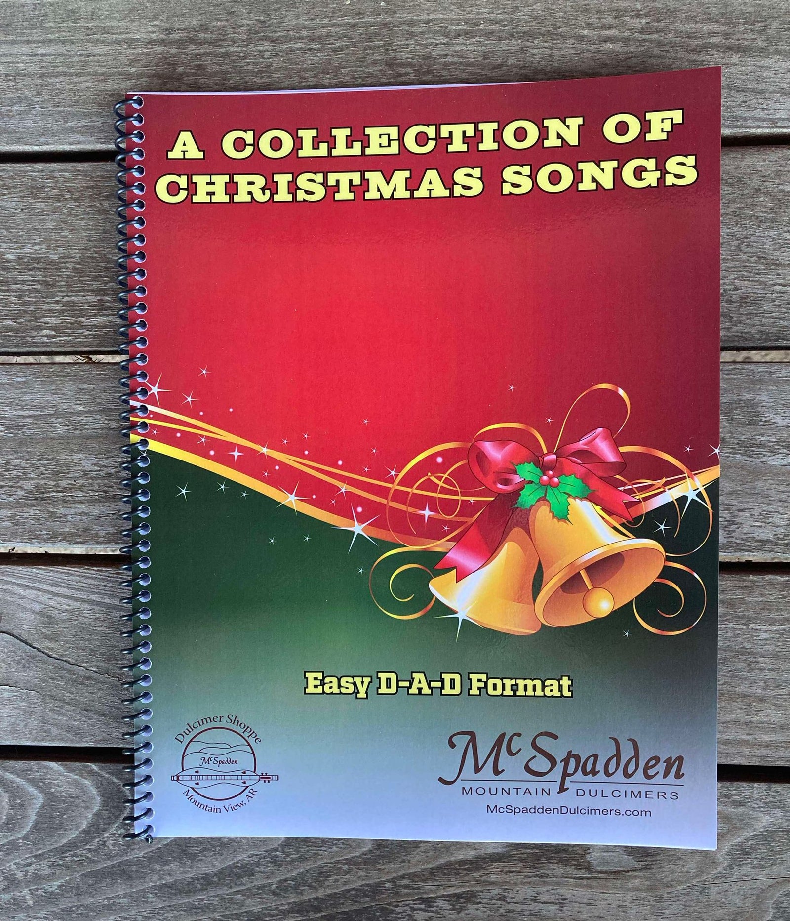 A spiral-bound book called "A Collection of Christmas Songs by Red Dog Jam (Giger)," ideal for mountain dulcimer fans. The cover showcases festive bell and ribbon graphics, bringing holiday cheer to your musical journey. Produced by McSpadden Mountain Dulcimers, this book caters to enthusiasts of the easy D-A-D format.