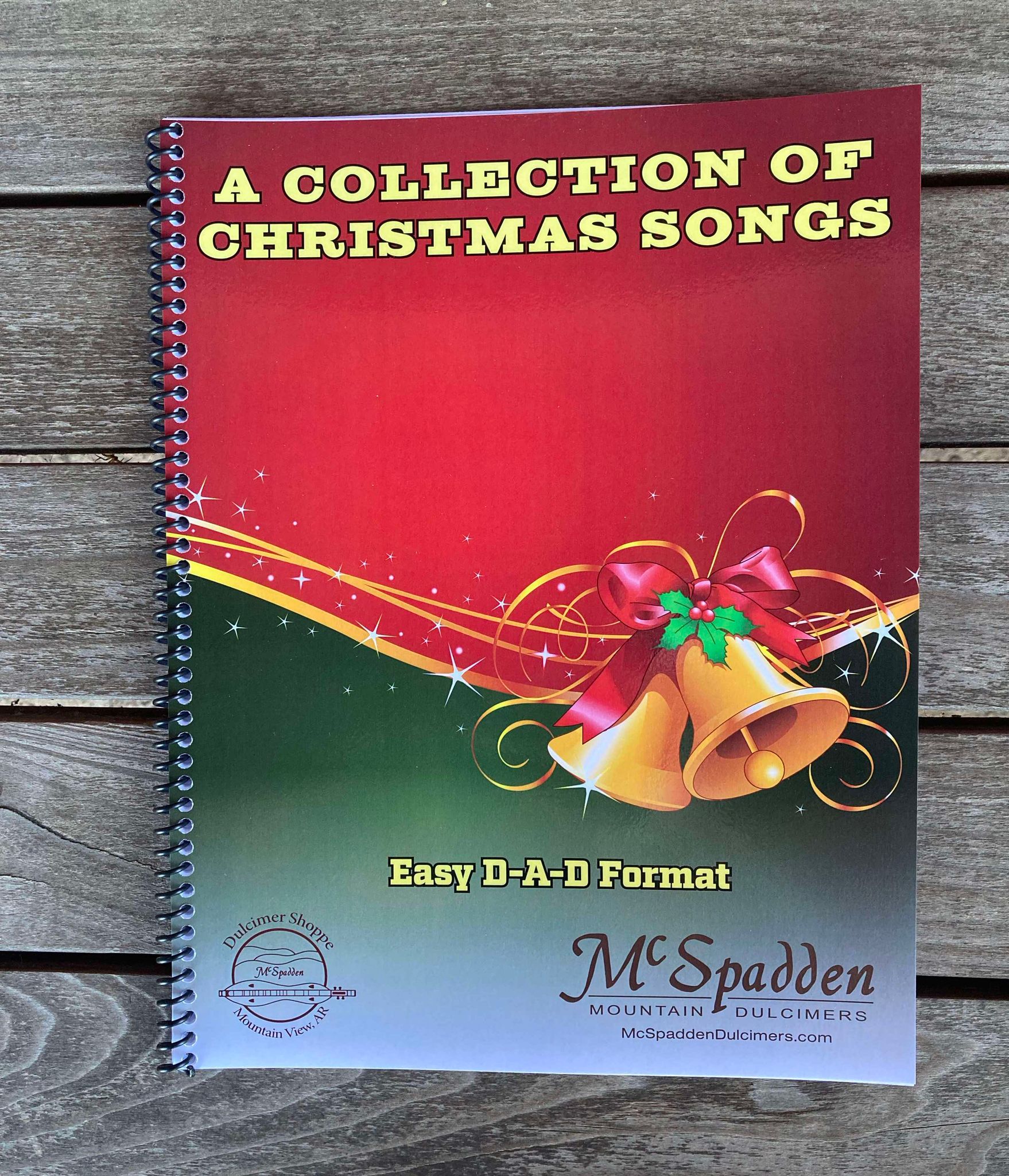 Spiral-bound booklet titled "A Collection of Christmas Songs by Red Dog Jam," featuring a red background with holiday bells and a bow, and subtitled "Easy D-A-D Format." Perfect for the mountain dulcimer enthusiast, this collection is published by McSpadden Mountain Dulcimers.