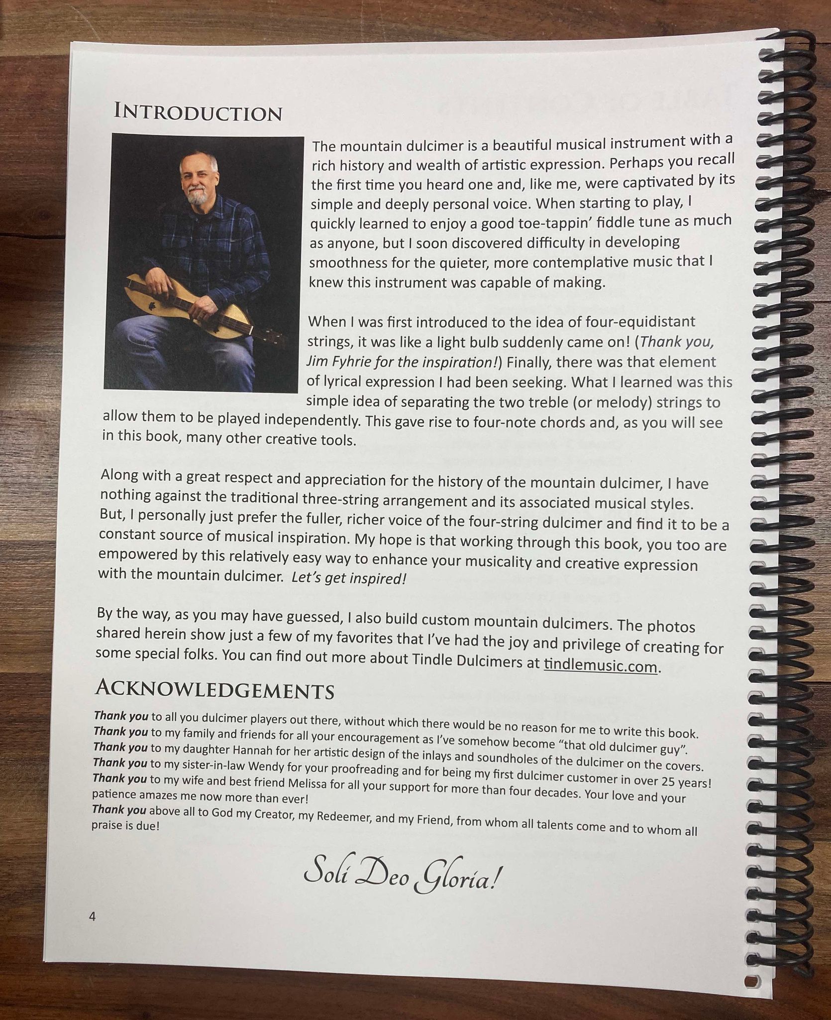 A spiral-bound book titled "The Essential Four String Dulcimer by Mark Tindle" open to a page labeled "Introduction," showcasing text about the mountain dulcimer, its history, and the author's experience with D-A-D-D tuning. In the top left corner, there is a picture of a man holding the four-string dulcimer.