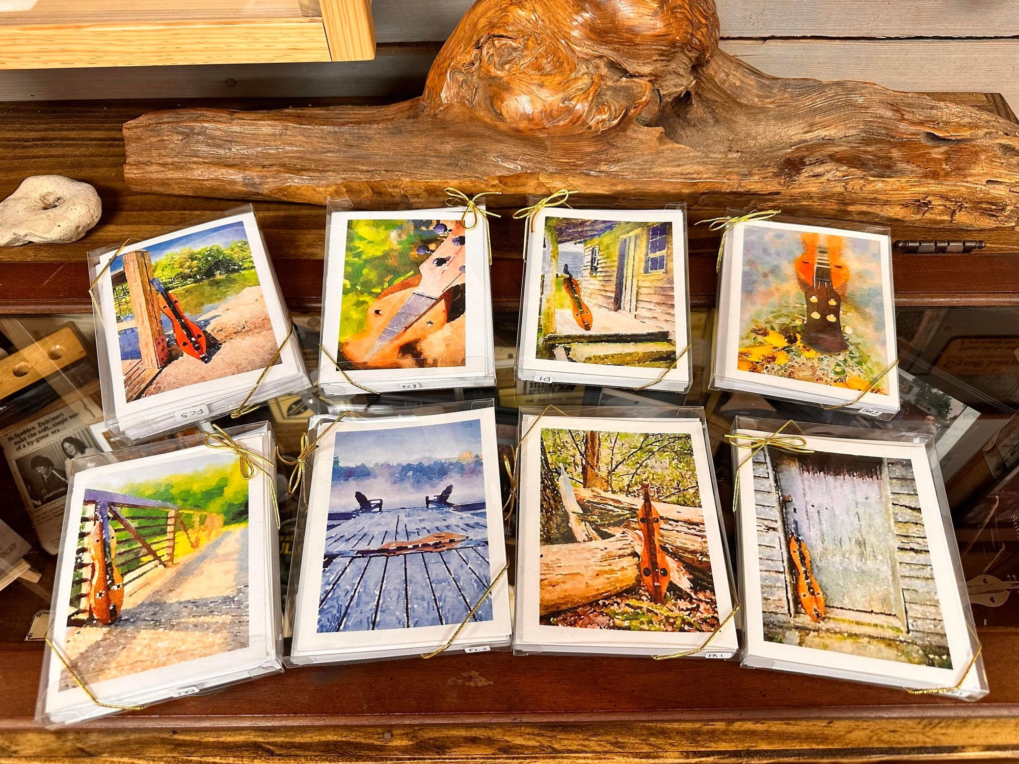 Eight photo cards featuring acoustic instruments in rustic outdoor settings are displayed against a wooden background, tied together with gold strings. These charming pieces evoke the essence of Dulcimer Greeting Card Sets by Ellen Pratt.