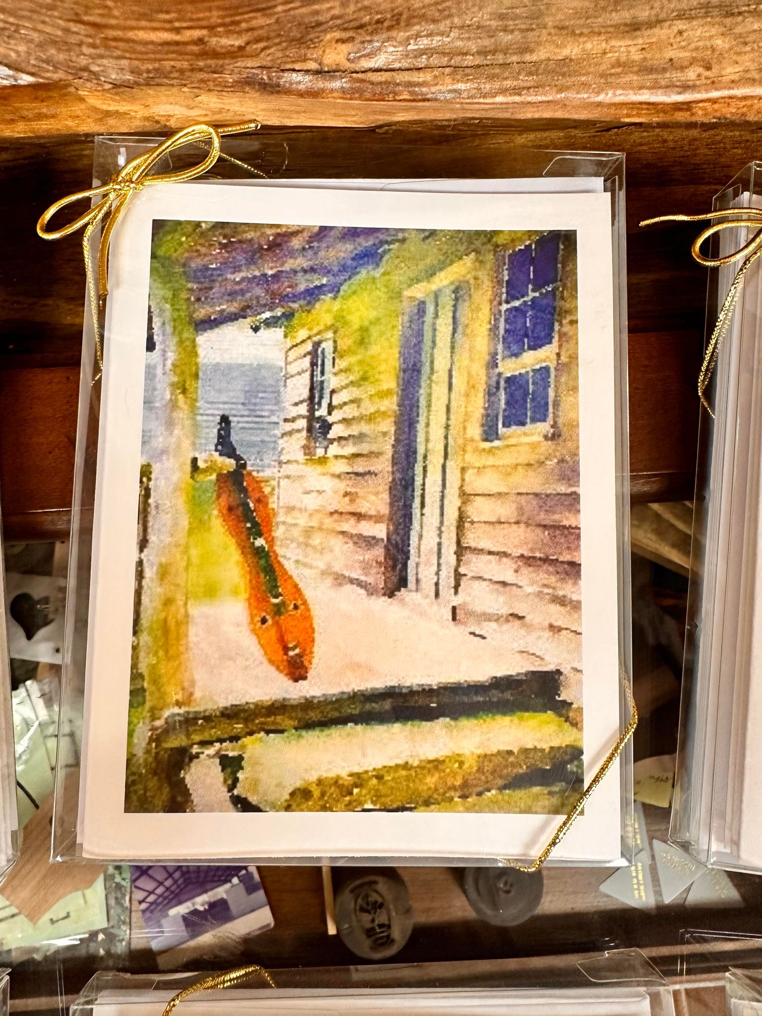 A photo of an artwork by Ellen Pratt depicting a skateboard leaning against the exterior wall of a building with beige siding, a window, and a door. The Dulcimer Greeting Card Sets by Ellen Pratt are wrapped in plastic with golden ribbon ties, reminiscent of the charm found in hand drawn greeting cards.