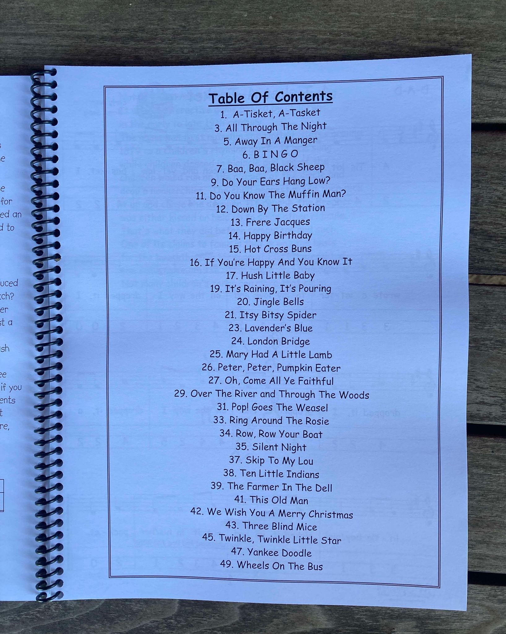 Table of contents of a beginner's book for nursery rhymes, featuring titles like "A-Tisket, A-Tasket," "Do You Know The Muffin Man?" and "The Ants Go Marching." The page, designed with Grandpa's Musical Guide Book by Red Dog Jam, is spiral-bound and laid on a wooden surface.