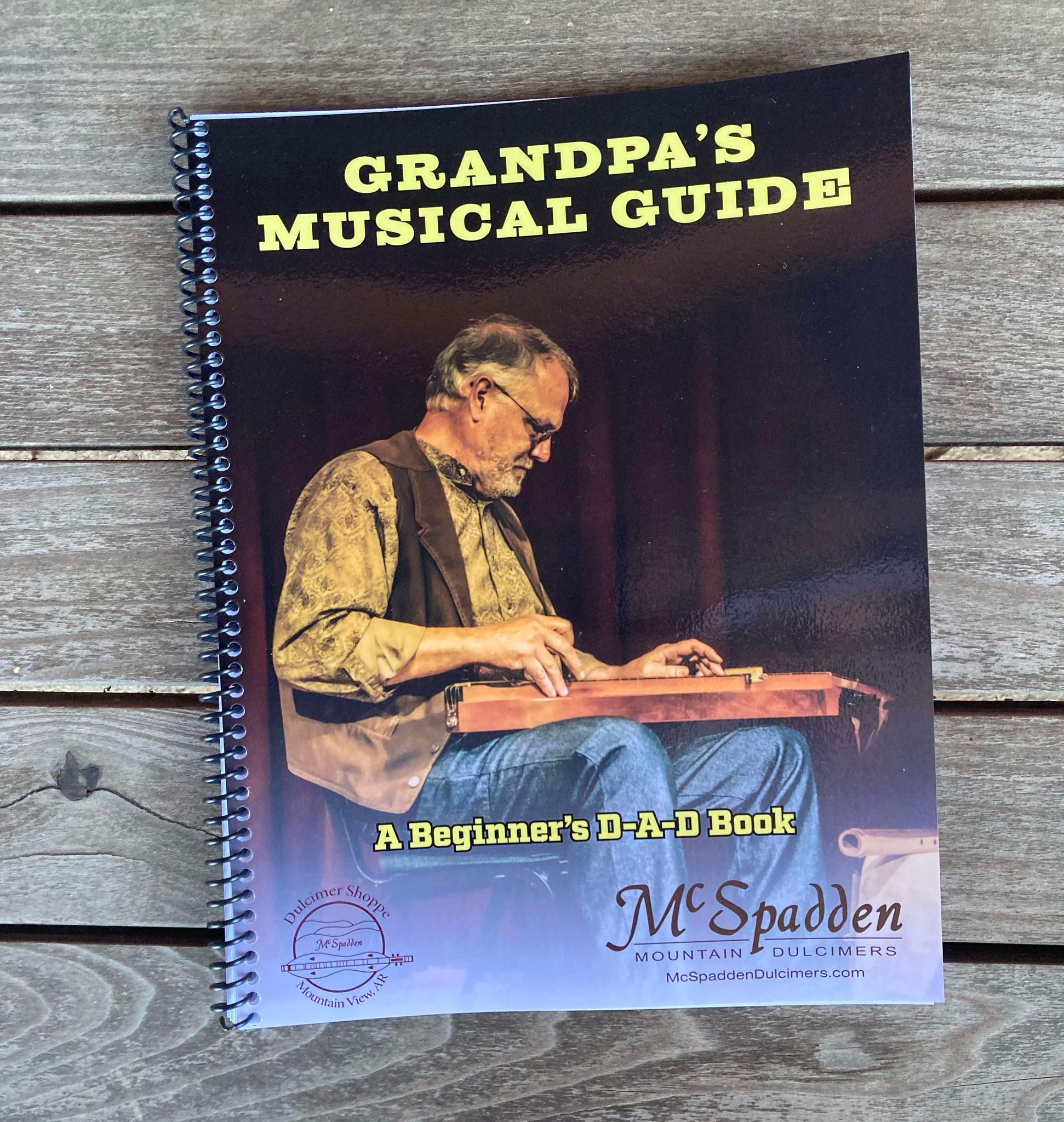 Cover of "Grandpa's Musical Guide or Songs Your Grandkids Should Know Book - by Red Dog Jam" featuring a man playing a mountain dulcimer, produced by McSpadden Mountain Dulcimers. The beginner's book is spiral-bound and rests on a wooden surface, ready to introduce you to classic nursery rhymes.