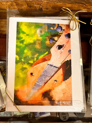 A framed watercolor painting of a hand-drawn stringed musical instrument by Ellen Pratt, set against an earthy, abstract background. The frame is tied with a gold ribbon and evokes the charm of Dulcimer Greeting Card Sets by Ellen Pratt.