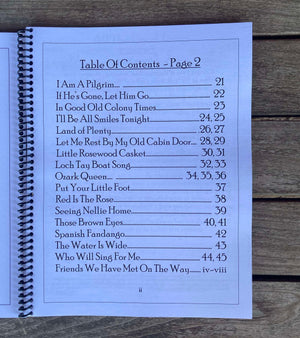 A spiral-bound book opened to a table of contents titled "Mountain View Treasures - by Red Dog Jam." It lists song titles, each with corresponding page numbers, labeled "Table Of Contents - Page 2." The book rests on a wooden surface, hinting at melodies played on a Mountain Dulcimer.