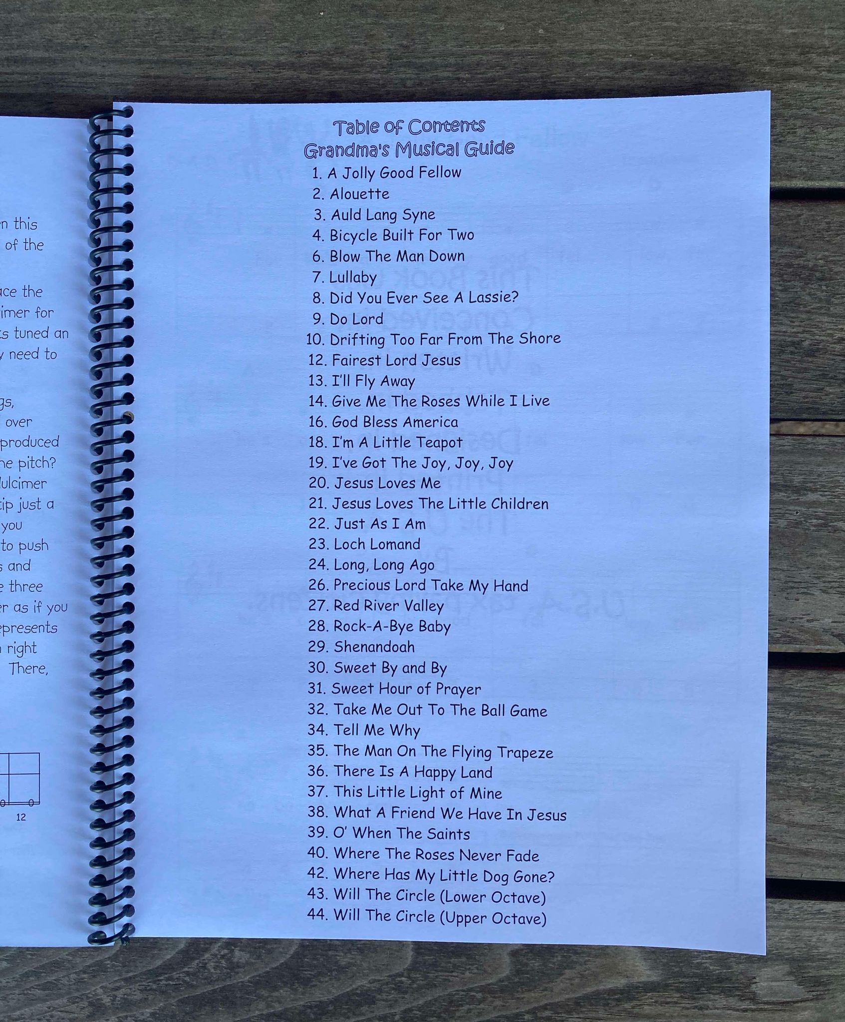A spiral-bound booklet opened to a table of contents titled “Grandma's Musical Guide - by Red Dog Jam,” listing song titles such as "Bicycle Built for Two," "Give Me the Roses While I Live," and "Will the Circle Be Unbroken?". This beginner's book even includes Mountain Dulcimer Tab for D-A-D tuning.