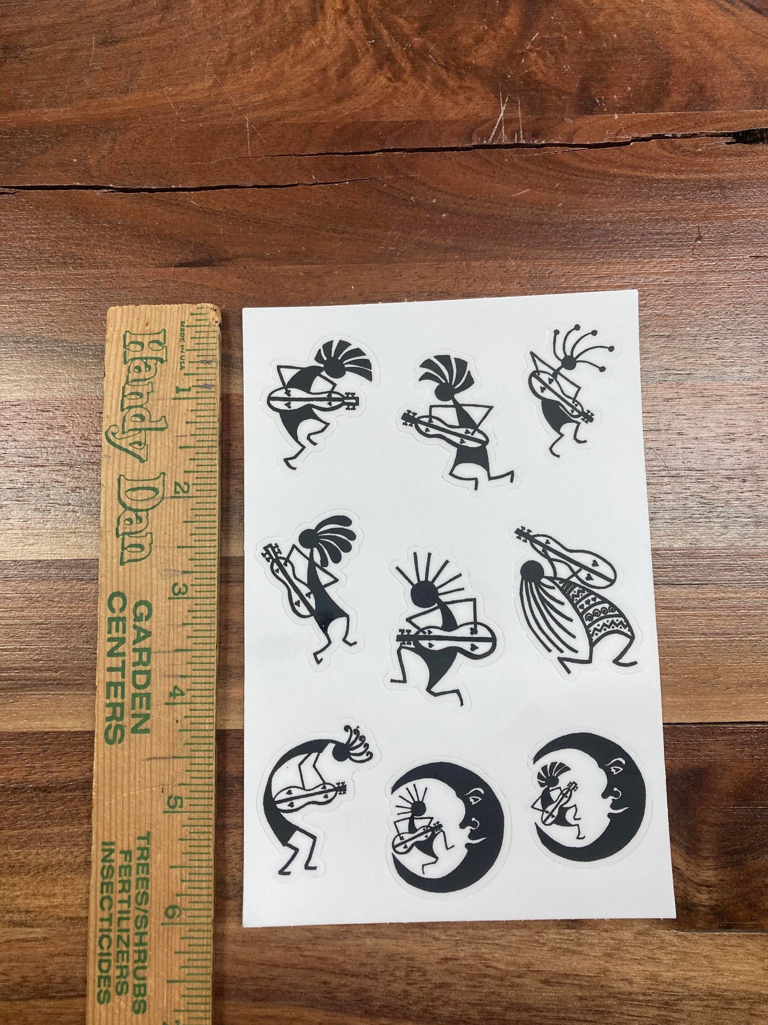 A sheet of McSpadden Stickers with various Kokopelli figures in black on white is placed on a wooden surface next to a wooden ruler, perfect for decorating and spreading joy.